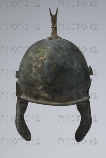 Bronze helmet, 4th–3rd century BC, Italy, armament; height 34 cm, width 20.2 cm, depth 22.5 cm, weight 1.035 kg, An aged metal helmet with a rounded dome and a small upward forked finial on top, showing a mottled surface of green, dark green, brown, and dark gray patina; a brim that follows the dome’s curve and a pair of curved cheek guards hanging down on either side in similar dark gray and brown tones; small rectangular attachments at the sides where the cheek guards connect; an overall weathered, textured appearance with patches of lighter green and brown across the surface against a pale gray background.