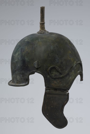 Bronze helmet, 4th–3rd century BC, Italy, armament; height 34 cm, width 20.2 cm, depth 22.5 cm, weight 1.035 kg., An aged metal helmet with a rounded dome and a short vertical spike rising from the top, showing a mottled surface of dark green, brown, and black patina; the helmet features a wide curved opening at the front and a hinged cheek guard hanging down on one side, both sharing the same dark greenish-brown coloration, with subtle areas of lighter green and yellowish discoloration and worn edges.