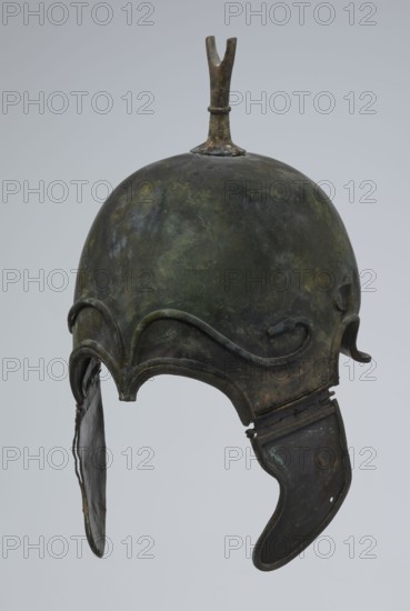 Bronze helmet, 4th–3rd century BC, Italy, armament; height 34 cm, width 20.2 cm, depth 22.5 cm, weight 1.035 kg., A metal helmet with a rounded dome and a small forked projection on top, showing a mottled surface of green, dark green, brown, and black patina; decorative raised curving ridges around the brow and cheek areas; a hinged cheek guard on one side with a curved lower edge and a hanging flap on the other side with a thin strip connecting it; overall worn and aged texture with uneven coloration against a pale gray background.
