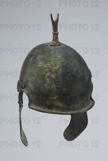 Bronze helmet, 4th–3rd century BC, Italy, armament; height 34 cm, width 20.2 cm, depth 22.5 cm, weight 1.035 kg, An aged metal helmet with a rounded dome showing mottled green, dark gray, brown, and patches of lighter green patina, topped by a vertical forked crest of darker bronze; a curved brim around the lower edge and a dangling ear flap on one side and a longer curved cheek guard on the other, both showing similar green and brown tones against a pale gray background.