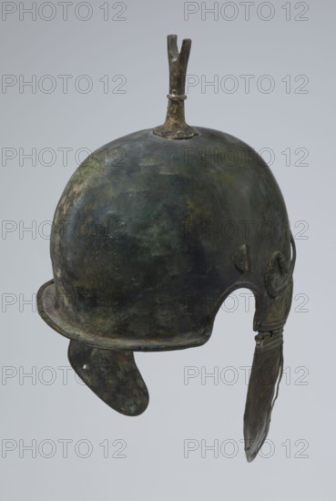 Bronze helmet, 4th–3rd century BC, Italy, armament; height 34 cm, width 20.2 cm, depth 22.5 cm, weight 1.035 kg, A worn metal helmet with a rounded dome and a short upright crest at the top, showing a mottled surface in dark green, bronze, brown, and hints of black; the helmet has a flared rim and a cutaway arch for the face, with a dangling cheek piece on one side and a curved protective flap on the other, both showing similar aged greenish-bronze and brown patina.