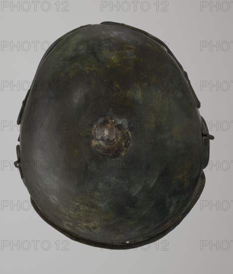 Bronze helmet, 4th–3rd century BC, Italy, armament; height 34 cm, width 20.2 cm, depth 22.5 cm, weight 1.035 kg., A domed metallic object viewed from above with an uneven, pitted surface showing a mix of dark green, brown, black, and hints of yellowish-green patina; a raised circular boss near the center with rough, corroded texture; the rim around the base is visible and shows darker brown and black tones; a few small attached metal fittings project from the sides, appearing darker and slightly rusted; the background is a uniform light gray.