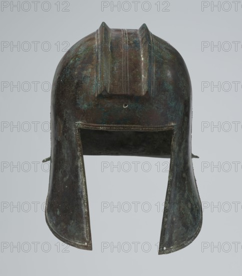 Bronze helmet, circa 550–500 BC, possibly Italy; armament. Dimensions: height 24 cm, width 18.5 cm, depth 23.2 cm; weight 1.170 kg., A single metal helmet viewed from the front with a rounded top and a raised central crest, showing a patina of greenish-blue and dark brown on its surface; a rectangular front opening leads into a darker interior, and two flared cheek guards extend downward on either side, each edged with a beaded trim and showing areas of mottled green-blue and brown; the background is a plain pale gray.