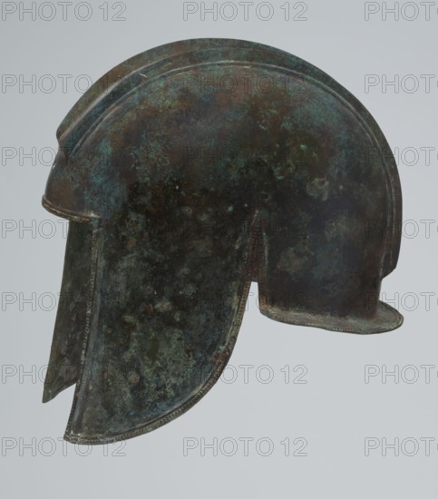 Bronze helmet, 550–500 BC, Italy, armament; height 24 cm, width 18.5 cm, depth 23.2 cm, weight 1.170 kg, An aged metal helmet shown in profile with a rounded dome and flared cheek guards, exhibiting a weathered surface mottled with dark brown, green, teal, and hints of rust-colored patina, with textured spots and areas of verdigris across the curved surfaces and edges.
