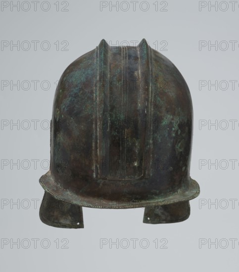 Bronze helmet, 550–500 BC, likely Italy, armament; height 24 cm, width 18.5 cm, depth 23.2 cm, weight 1.170 kg, A worn metal helmet shown from the front with a rounded dome and a short brim, displaying a mottled surface of dark brown and green patina with areas of bluish green corrosion; the helmet has raised ridges running from front to back and two earflaps hanging down at the sides, both showing similar brown and green discoloration and some surface roughness.