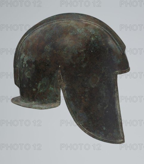 Bronze helmet, 550–500 BC, Italy, armament; height 24 cm, width 18.5 cm, depth 23.2 cm, weight 1.170 kg, A single metal helmet shown in profile against a pale gray background, with a rounded dome, a projecting brim at the back, and an elongated cheek guard at the front; the surface shows a mottled patina with dark brown, green, bluish-green, and black areas, and small patches of lighter green corrosion, with visible texture and slight irregularities along the rim and edges.