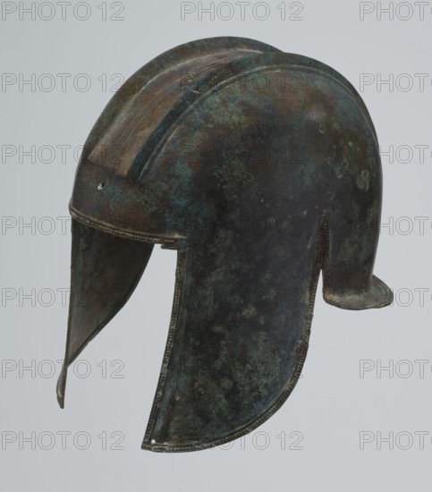 Bronze helmet, 550–500 BC, Italy, armament; height 24 cm, width 18.5 cm, depth 23.2 cm, weight 1.170 kg, A weathered metal helmet with a rounded dome and a pronounced central ridge, showing a mottled surface of green, brown, and dark bluish tones; the helmet has a forward-sloping brow and extended cheek guards with rough edges, a short neck guard at the back, and visible seams or rivet lines along the borders, with areas of darker patina and lighter greenish corrosion creating a textured appearance.