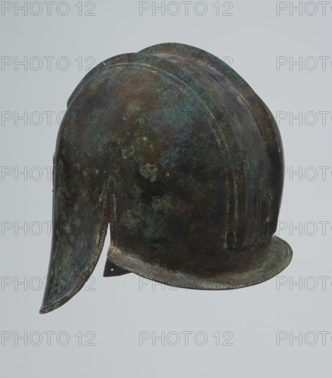 Bronze helmet, 550–500 BC, Italy, armament; height 24 cm, width 18.5 cm, depth 23.2 cm, weight 1.170 kg, A weathered metal helmet rests against a pale gray background, showing a rounded crown with a curved brim and an extended cheek guard on one side; the surface displays mottled patches of greenish patina, dark brown oxidation, and areas of bluish and black discoloration, with subtle ridges and beaten metal textures visible along the dome and brim.