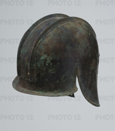 Bronze helmet, 550–500 BC, Italy, armament, height 24 cm, width 18.5 cm, depth 23.2 cm, weight 1.170 kg, A metal helmet with a rounded dome and a rear neck guard, showing a mottled surface of green, brown, dark bluish, and black tones, with areas of lighter green patina and darker brown spots, and a curved brim along the front edge.