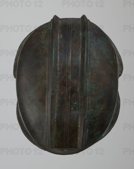 Bronze helmet, 550–500 BC, Italy, armament; height 24 cm, width 18.5 cm, depth 23.2 cm, weight 1.170 kg, A close-up view of a rounded metallic object with a symmetrical ridged central spine and raised edges, showing a weathered surface with patina. The object displays a mix of dark brown, green, bluish-green, and hints of black tones, with areas of mottled discoloration and subtle reflective highlights. The background is a uniform light gray.