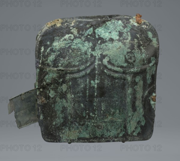 Two-piece torso armor with buckles, 4th century BC 375–325 BC, Southern Italy, weaponry; maker, two-piece cuirass design., An aged metal object with a rectangular rounded shape sits against a neutral gray background, showing a patina of green and teal corrosion mottled over dark gray and almost black areas; the surface has irregular patches of lighter green, turquoise, and pale mint along with small rusty brown spots and flaking where the metal has worn away, including a rust-colored crust near the top edge and a torn, bent tab protruding from one side that shows similar green and gray tones; the front displays subtle raised curved ridges and shallow indentations, and the edges are worn and uneven with small holes and rough textures.