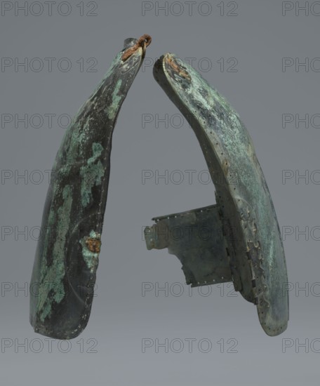 Two-piece torso armor with buckles, 4th century BC 375–325 BC, Southern Italy, weaponry., A pair of curved, weathered metal pieces positioned facing each other against a plain gray background, each showing patches of greenish patina and darker areas of black and brown, with a few rusty orange spots and traces of rough texture; one piece has a small rusted loop at its top edge and the other shows a jagged interior plate attached near its lower middle, with several evenly spaced small holes along one edge and irregular edges and worn surfaces throughout.