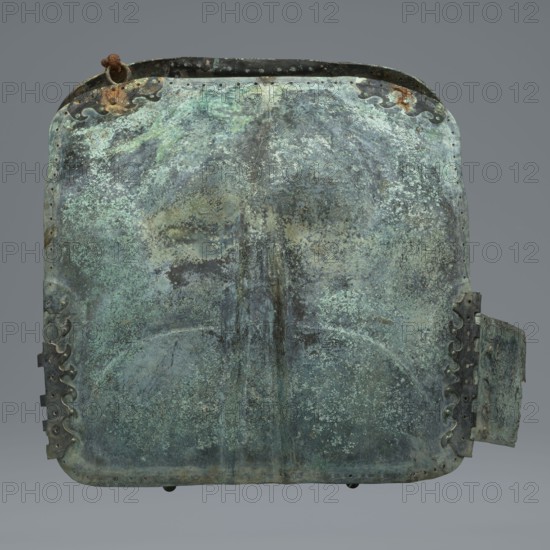 Two-piece torso armor with buckles, 4th century BC 375–325 BC, Southern Italy ?, weaponry., A large rectangular metal object with rounded corners fills the frame, showing a heavily weathered surface in shades of green, teal, blue-green, dark gray, and patches of brownish rust; the metal has a mottled, pitted texture with areas of lighter greenish corrosion and darker worn spots, and a faint central vertical ridge running from top to bottom; along the top and sides there are ornamental wavy metal strips and rivet holes, and a rusted metal loop is attached near the upper left edge; a hinged or protruding panel is visible on the right side with similar corrosion and decorative edging.