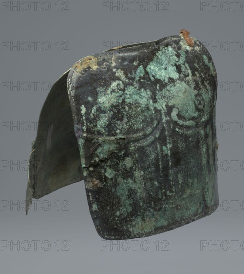 Two-piece torso armor with buckles, 4th century BC 375–325 BC, Southern Italy, weaponry, An aged metal helmet is shown against a plain gray background, with a patinated surface displaying shades of green and turquoise mixed with dark oxidized black and brown. The helmet has a rounded top and a flared lower edge, with visible irregular mottling and corrosion patterns across its surface. A few areas show rusty orange-brown spots and worn metallic edges, and a group of small perforations lines the upper rim. The interior visible at the side is darker with greenish and brown tones, and the overall surface texture appears rough and pitted with areas of flaking and uneven coloration.