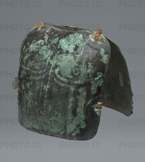 Two-piece torso armor with buckles, 4th century BC 375–325 BC, possibly Southern Italy; weaponry., An aged metal helmet with a rounded top and a flared back, showing an uneven surface covered in mottled patches of green and dark brownish-black patina, with areas of lighter green corrosion and small rusty brown encrustations near seams; the metal has visible rivet lines and worn edges, and the interior visible at the rear appears darker with greenish tones, all set against a plain gray background.
