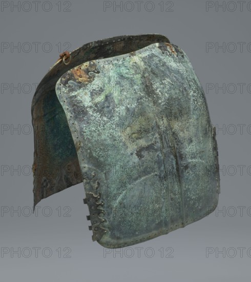 Two-piece torso armor with buckles, 4th century BC 375–325 BC; Southern Italy ?, weaponry., An aged metal helmet with a curved, rounded form sits against a plain gray background; its surface shows a mottled blend of greenish-blue patina, pale green, dark teal, and areas of brownish rust, with patches of darker gray and black; decorative scalloped edges are visible along the front and sides, and a small rusty ring with a short cord is attached near the top; the interior surface appears darker with green and brown corrosion, and the metal shows indentations, rivet holes, and worn textures across the exterior.