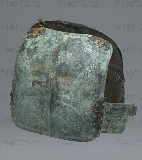 Two-piece torso armor with buckles, 4th century BC 375–325 BC, Southern Italy, weaponry, A corroded metal chestplate with a mottled surface showing shades of green, turquoise, pale blue, dark gray, and brown, with areas of rust and darker brown oxidation; the plate has curved edges, rivet holes along the borders, decorative scalloped metal fittings with darker patina, a hinge-like attachment on one side, and a small rusty ring near the top edge; the inner surface visible at the opening appears darker with rough, rusty brown and dark green textures, all shown against a neutral gray background.