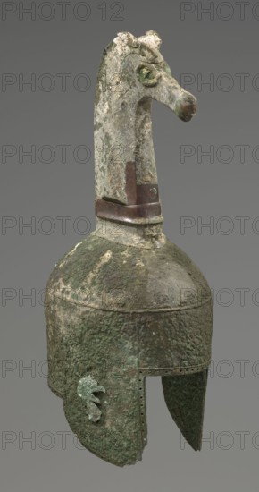 Bronze helmet, 8th–7th century BC, Italy, armament; height 46 cm, width 17.5 cm, depth 23 cm, weight 2.740 kg., An aged metal helmet with a long neck crest shaped like an animal head protruding upward, showing rough, corroded surfaces in shades of green, brown, and gray; the helmet body is rounded with a textured, pitted surface and a patina of green and darker brown, with a few lighter patches, and a band near the base of the crest that appears darker brown; the lower edge of the helmet has a cutaway face opening with a row of small rivet holes along the rim and a decorative raised element on one side, all exhibiting weathered green and brown corrosion against a neutral gray background.