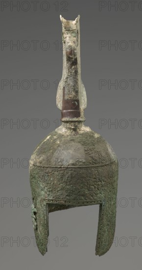 Bronze helmet, 8th–7th century BC, Italy, armament; height 46 cm, width 17.5 cm, depth 23 cm, weight 2.740 kg, A weathered metallic object with a rounded dome and elongated cheek guards, showing a rough, pitted surface texture and areas of corrosion; the top features a vertical crest or ornament resembling an animal head with a pronounced snout and flared ears, attached to a short neck above the dome; the metal shows a range of colors including greenish patina, brownish and dark brown patches, and some lighter gray and off-white encrustations; the open lower edge of the helmet has a rectangular face opening and small perforations along the rim, and the whole piece rests against a plain neutral gray background.