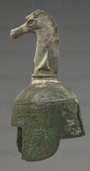Bronze helmet, 8th–7th century BC, Italy, armament; height 46 cm, width 17.5 cm, depth 23 cm, weight 2.740 kg., A weathered metallic helmet with a textured, pitted surface and a greenish patina, topped by a sculpted animal head crest that resembles a long-necked creature with a pronounced snout and carved eye. The helmet shows areas of darker green and brown oxidation, with lighter patches of exposed metal where the surface is eroded. The crest and helmet display rough, uneven textures and flaking, and the helmet has a downward-curving face guard with a row of small perforations along its edge. The background is a uniform gray.