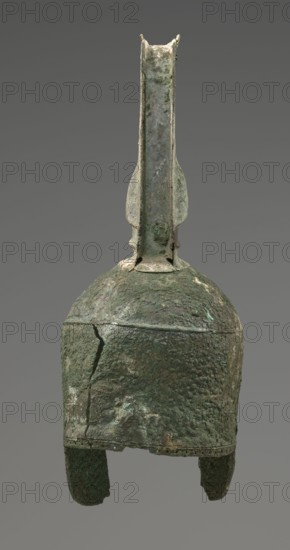 Bronze helmet, 8th/7th century BC, Italy, armament; height 46 cm, width 17.5 cm, depth 23 cm, weight 2.740 kg., An aged metal helmet with a tall central crest and a rounded dome-shaped top, showing a rough, pitted surface and visible cracks; the helmet has two short cheek pieces extending downward and a vertical fin-like ridge rising from the crown; colors visible include various shades of green, dark green, brown, gray, and traces of lighter green patina against a neutral gray background.