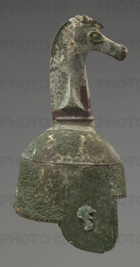 Bronze helmet, 8th/7th century BC, Italy, armament; height 46 cm, width 17.5 cm, depth 23 cm, weight 2.740 kg, The image shows an aged metal helmet with a long neckpiece topped by a stylized animal head. The surface of the helmet and crest is heavily corroded and pitted, showing a mix of green, brown, gray, and dark green patina with areas of lighter and darker mottling. The animal head has a rounded snout and a raised eye area with similar corrosion and color variation, and a darker band around the base of the crest. The dome of the helmet is rounded and textured with rough corrosion, and the attached cheek guard hangs down with an irregular edge and a small attached decorative piece that is a lighter green compared with the surrounding darker corrosion. The background is a smooth neutral gray.