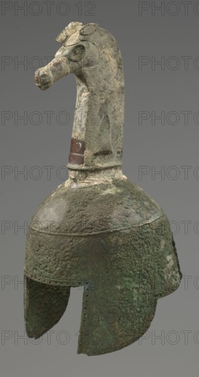 Bronze helmet, 8th–7th century BC, Italy, armament, height 46 cm, width 17.5 cm, depth 23 cm, weight 2.740 kg, A weathered metal helmet with a long neck crest shaped like a stylized animal head rises from the dome; the crest has a textured, pitted surface and traces of lighter and darker areas. The helmet body is rounded with flared cheek guards and shows heavy corrosion and rough, bubbly texture. Visible colors include greenish patina, dark brown, dull gray, and patches of lighter beige and reddish-brown on the crest. The background is a uniform gray.
