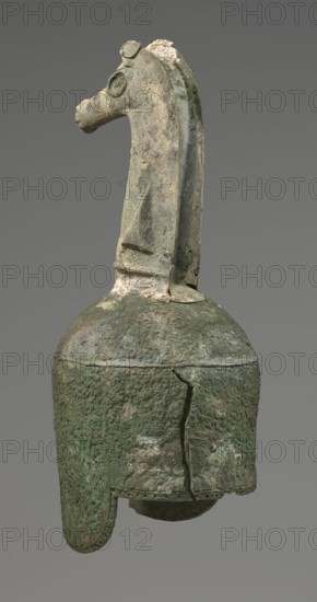 Bronze helmet, 8th/7th century BC, Italy, armament; height 46 cm, width 17.5 cm, depth 23 cm, weight 2.740 kg., A weathered sculpted object resembling a long-necked animal head sits atop a rounded, bell-like base; the surface shows rough, pitted texture and areas of encrustation. The neck and head feature a raised crest and a circular eye shape, with a muzzle projecting forward. The bell-like lower portion has a flared rim and a visible crack running down its side, and a rounded inner element is partly visible beneath the rim. The visible colors are shades of green, muted gray, pale beige, and hints of brown.