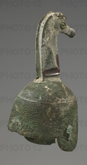 Bronze helmet, 8th–7th century BC, Italy, armament; height 46 cm, width 17.5 cm, depth 23 cm, weight 2.740 kg;, The image shows a weathered metal object resembling a helmet with a long decorative crest shaped like a stylized animal head. The surface displays a patina in shades of green and dark green with areas of brown and gray, and the metal texture appears rough and pitted. The crest curves upward and forward, forming an animal-like head with a pronounced snout and a rounded eye area. The main dome of the helmet is rounded with an extended neck guard and ear-covering flaps, all showing uneven corrosion and encrustation. There are subtle variations of lighter green and pale discoloration across the surface, and a narrow darker band is visible near the base of the crest. The background is a uniform mid-gray.
