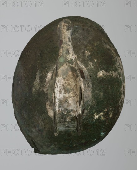 Bronze helmet, 8th/7th century BC, Italy, armament; maker, height 46 cm, width 17.5 cm, depth 23 cm, weight 2.740 kg, An oval object with a weathered surface showing a mix of dark green, brown, beige, and pale off-white areas, featuring a central raised ridge or handle-like protrusion that is lighter in color with patches of green and beige, and several irregular rough textures and mottled discolored spots across the surface.
