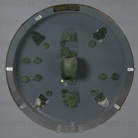 Hoplite shield, 6th century BC, Italy, weaponry, A round display with a muted gray background holds a group of greenish metal fragments arranged symmetrically around a central cylindrical object; the metal pieces show varied green patina and some darker verdigris, with a few fragments that include areas of silvery metal. The central cylindrical piece is green with mottled darker spots. Around it are several decorative pieces with scalloped, floral, and fan-like shapes in green tones, some attached to small round green discs. The display rim is metallic with a darker outer edge and small metal brackets, showing brownish corrosion in places; the overall colors visible are shades of gray, green, dark green, silver, brown, and a small circular spot of gold-toned color.