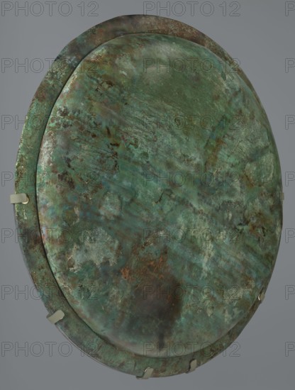Hoplite shield, 6th century BC, Italy, weaponry, creator unknown, maker: Guttmann, Axel, Scen.Gal.Klass., A large roundish metal object with a weathered, pitted surface showing a mix of green and turquoise patina with areas of dark brown and black discoloration, lighter green and blue-green mottling, and some pale grayish patches; the rim appears slightly raised and shares the same varied green and brown corrosion tones, and a few small pale beige metal clips are attached around the edge against a plain neutral gray background.