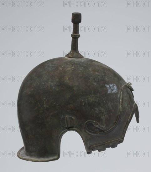 Bronze helmet, 4th–3rd century BC, Italy, armament; height 26.5 cm without cheek guards, width 19.8 cm, depth 21.6 cm, weight 0.758 kg without cheek guards., An aged metallic helmet with a rounded dome shape and a short brim at the back, showing a patina of dark brown, greenish, and muted bluish tones, with a vertical finlike knob rising from the top that is darker brown, and faint lighter spots on the dome; the helmet has a curved cheek guard on one side with an attached decorative curling strip and a small ring at its upper edge, and a semicircular cutout at the bottom edge.