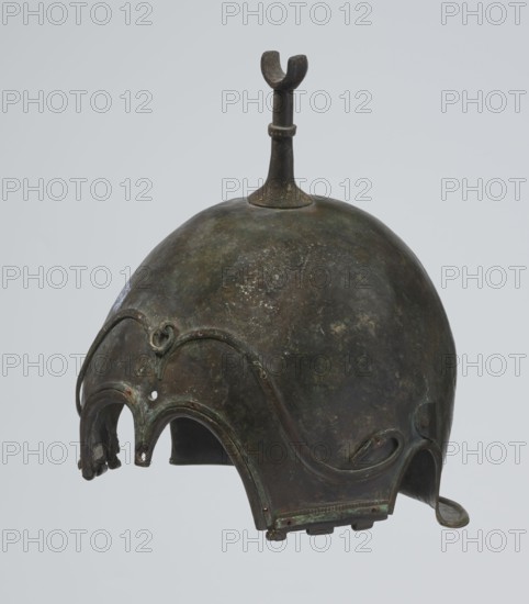 Bronze helmet, 4th–3rd century BC, Italy, armament; height 26.5 cm without cheek guards, width 19.8 cm, depth 21.6 cm, weight 0.758 kg without cheek guards., A rounded metal helmet with a domed top and a vertical fin-like projection rising from its crown, the surface showing mottled dark green, brown, and black patina with areas of lighter green corrosion; the front edge has an ornate scalloped rim with a few arching cutouts and curved decorative ridges, including a small raised loop detail near the forehead; the lower edges feature attached flaps and tabs with worn greenish and brown tones and a small hooked extension on one side; the overall texture appears aged and uneven with patches of darker and lighter coloration against a plain light gray background.