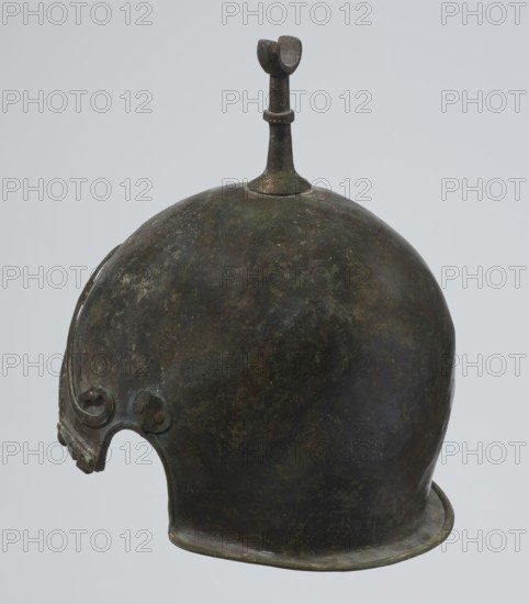Bronze helmet, 4th–3rd century BC, Italy, armament, height 26.5 cm without cheek guards, width 19.8 cm, depth 21.6 cm, weight 0.758 kg without cheek guards, A rounded metal helmet with a patinated surface showing shades of dark green, brown, and black, featuring a raised knob and short vertical fin at the top that is darker and slightly textured, a curved brim along the lower edge, and a decorative cutaway at the front with a small attached hinge-like element and subtle ornamental ridges; the background is a light gray.