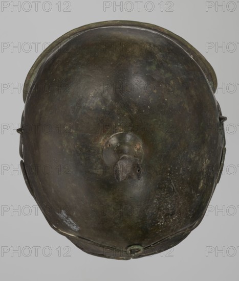 Bronze helmet, 4th–3rd century BC, Italy, armament; height 26.5 cm without cheek guards, width 19.8 cm, depth 21.6 cm, weight 0.758 kg without cheek guards., A top-down view of a rounded metal object with a smooth domed surface showing a patina of dark brown, green, and hints of black, with areas of lighter greenish corrosion and small pale speckles; a raised circular boss sits near the center with a short protruding stub that appears darker and rougher; the rim around the object is slightly raised and shows similar brown and green tones, and a few small attachments or fittings are visible along the sides and lower edge in matching dark metal colors.
