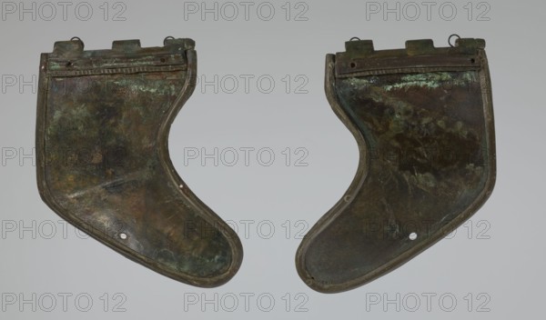 Bronze helmet, 4th–3rd century BC, Italy, armament; height 26.5 cm without cheek guards, width 19.8 cm, depth 21.6 cm, weight 0.758 kg without cheek guards, A pair of curved metal plates displayed against a pale gray background, each plate showing a weathered, mottled surface with dark brown and deep green patina and areas of bluish-green corrosion; the plates have rounded outer edges and squared upper sections with small looped fittings, and each contains a small circular hole near the lower curved area; the overall tones include brown, green, bluish-green, and hints of black and dull metallic gray.
