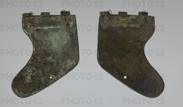 Bronze helmet, 4th–3rd century BC, Italy, armament; maker, height 26.5 cm without cheek guards, width 19.8 cm, depth 21.6 cm, weight 0.758 kg without cheek guards., A pair of irregularly shaped flat metal plates with curved lower edges and notched tops, shown side by side against a light gray background; the plates have a weathered, mottled surface with predominant greenish patina, dark brown and rusty orange areas, and touches of bluish-green and black; each plate has small circular holes near the lower portion and small metal loops or rings attached along the top edge; the surfaces show uneven textures, patches of corrosion, and variations in color intensity.