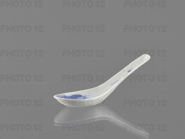 Spoon, bats iconography, ceramics, Harbin China, figurative scenes, blue and white porcelain, Qing style, Chinese art/culture, cutlery, Qing 1644–1911, 18th century, China, length 12.5 cm, A ceramic spoon with a curved handle rests on a reflective surface, showing a mirror-like reflection beneath it; the spoon is primarily white with a few blue brushlike marks on the bowl and along the handle, and the background and surface are shades of gray.
