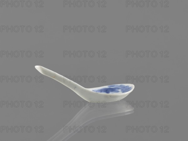 Spoon, bats iconography, ceramics, Harbin China, figurative scenes, blue-and-white porcelain, Qing style, Chinese art/culture, cutlery, Qing 1644–1911, 18th century, China, length 12.5 cm, A small ceramic spoon rests on a smooth reflective surface, showing a reflection beneath it; the spoon is predominantly white with a rounded handle and a shallow bowl that has blue brushlike patterns inside; the background and surface are various shades of gray, and the reflection shows muted tones of white and blue.