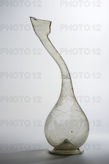 Bottle with a swan neck ashkdan, Islamic/Persian art, Persian culture, Qajar dynasty 1785–1925, 2nd half 18th–1st half 19th century, Iran; vessel utility glass, height 29.3 cm, width 13.0 cm, depth 10.0 cm. Characteristic swan neck with a separated spout; neck slightly profiled due to bending at the neck molding; used in everyday life in the Middle East., A translucent pale greenish-gray blown glass vessel with a rounded bulbous base tapering into a long, curved neck that ends in an irregular, angled lip, showing a few surface imperfections and internal bubbles, sitting on a flat circular foot against a light gray to white background.