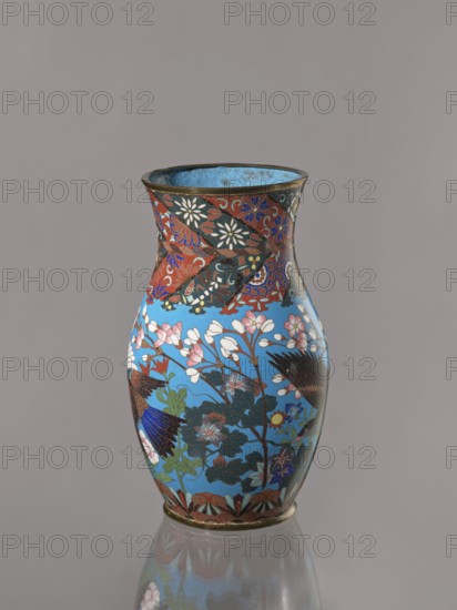 Vase, Revindication Kraków. Doves, flowers iconography; Japanese art; Japanese culture. Choynowski, Józef 1833–ca.1915. Birds. Meiji 1868–1912. Peonies, chrysanthemums, wolfberries, kachôga iconography, sakura iconography. Cloisonné enamel. Belly flowing smoothly into a short, thick, slightly parted neck. Cloisonné enamel on copper; brass foot and spout frame. Ca. 1870–1880 Meiji Period, 1868–1912. Japan, Nagoya. Vessel. Height 26.5 cm; width 12.6 cm., A decorative vase with a flared rim and rounded body sits against a plain gray background, featuring a glossy sky-blue base color covered with floral and bird motifs in a palette of red, white, pink, green, dark blue, black, and gold; the neck area shows a band of red and black patterned sections with white and blue floral accents, while the main body displays branches with white and pink blossoms, dark green leaves, and stylized birds with dark blue and black feathers, and a narrow gold rim and base.