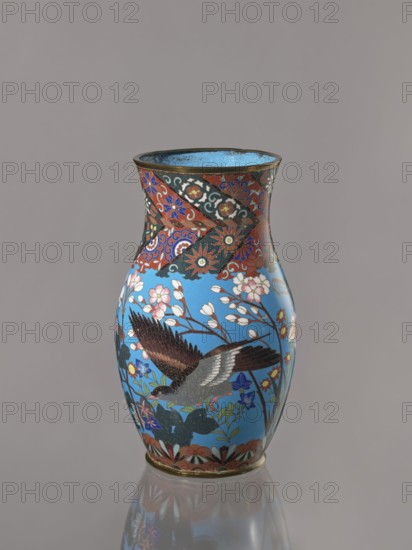 Vase, Doves, flowers iconography; Japanese art; Japanese culture; birds; Meiji 1868–1912; peonies; chrysanthemums; wolfberries; kachôga iconography; sakura iconography; cloisonné enamel. Belly flowing smoothly into a short, thick, slightly parted neck. Cloisonné enamel on copper; brass foot and spout frame. Ca. 1870–1880 Meiji Period, 1868–1912. Japan, Nagoya. Vessel; height 26.5 cm, width 12.6 cm., A decorative vase stands against a plain gray background, predominantly showing bright blue, red, black, white, brown, pink, green, and gold colors; its surface features floral motifs and stylized patterns with a large bird in flight depicted in shades of brown, black, gray, and white among branches with pink and white blossoms, surrounded by green leaves and various small flowers in blue, yellow, and red; the neck has ornate banded designs in red, black, blue, and gold, and the rim and base show metallic gold tones; the vase casts a soft reflection on the glossy surface beneath it.
