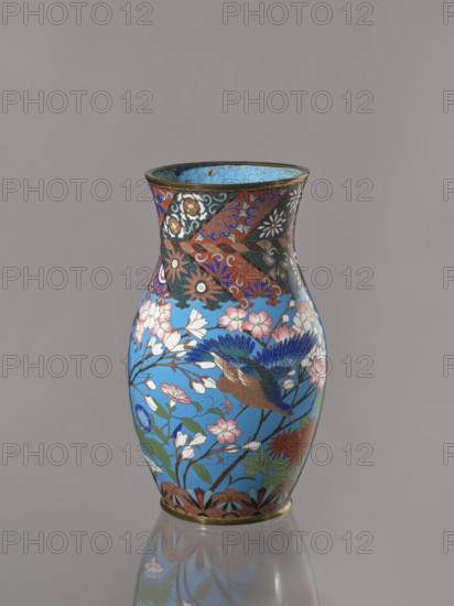 Vase, Doves, flowers, birds, lilies, chrysanthemums, wolfberries, sakura, butterflies, dragonflies — kachôga iconography. Japanese, Meiji Period ca. 1870–1880. Vase with a thick neck, parted towards the spout. Cloisonné enamel on copper; spout and foot enclosed by brass rings. Nagoya, Japan. Vessel; height 26.5 cm, width 15.5 cm., A tall, narrow vase with a flared rim sits against a plain light gray background, its surface covered in colorful floral and bird motifs. The main field of the vase is bright sky blue, decorated with a group of pink and white blossoms on thin brown branches and green leaves. A bird with deep blue wings, brown body, and darker feather details appears in flight among the flowers. The upper section of the vase features a band of intricate patterns in deep red, black, white, and blue with stylized floral shapes, while the lower rim shows a decorative border in muted brown and black with touches of blue. The interior of the rim is a lighter blue, and the vase rest...