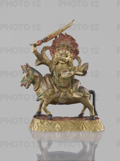 Palden Lhamo, Tibetan Buddhist figural sculpture dharmapala, 19th century, possibly China or Mongolia; materials not specified; depicts Palden Lhamo with mules and skulls; height 14.8 cm, width 13.0 cm., A small gilded metal figure of a fierce deity with a red mane of hair sits astride a horse, the deity holding a curved sword aloft in one hand and a shallow bowl in the other; the figure wears ornate armor and jewelry with traces of green and red paint, and the horse has painted green and red details on its bridle and mouth and a black tail and hooves. The sculpture stands on an oval decorative base with a repeating geometric pattern in gold and red, and the whole object is set against a plain light gray background with a faint reflection visible below.