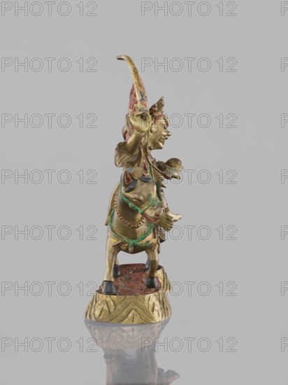 Palden Lhamo; unknown creator; Tibetan Buddhist art figurine; dharmapala iconography depicting mules and skulls; 19th century; China, or Mongolia ; sculpture; height 14.8 cm, width 13.0 cm., A small gilded figurine of a person riding an animal, shown in profile against a pale gray background; the figure and animal are mainly gold with accents of green, red, blue, black, and traces of darker brown, and they stand on a textured gold-colored base with a red surface area beneath the animal, the object casting a faint reflection below.