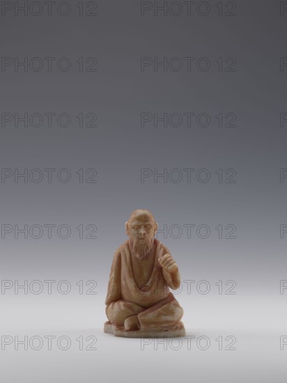 Sage, unknown sculptor, figurine of a bonza priest, 19th century, China; Chinese art; Qing 1644–1911; maker possibly Choynowski, Józef 1833–ca.1915 or Dawidow, Alexander Petrovich 1838–1885; sculpture; height 10.4 cm, width 6.9 cm, depth 5.0., A small carved figure of a seated bearded man in a robe, rendered in shades of warm tan and brown with lighter patches, is placed on a smooth pale surface against a gradient background that shifts from light gray near the bottom to darker gray toward the top. The figure sits cross-legged on a rounded base with one hand resting on the knee and the other raised slightly; facial features and a detailed beard are visible, and the robe drapes over the shoulders and legs creating folds. The overall colors visible are tan, brown, pale beige, light gray, and darker gray.