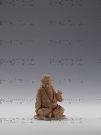 Sage, unknown sculptor, figurine of a bonza priest, Chinese, Qing dynasty 1644–1911, 19th century, stone sculpture, height 10.4 cm, width 6.9 cm, depth 5.0 cm; associated names: Choynowski, Józef 1833–ca.1915 and Dawidow, Alexander Petrovich 1838–1885., A small carved figurine of a seated elderly man with a long beard shown against a gradient gray background, the figure appearing in warm tones of beige, tan, and light brown with subtle variations and speckles, wearing draped robes and sitting cross-legged on a rounded base, one hand resting on his knee and the other raised with fingers curled, facial features and beard carved in modest detail, the surface showing slight mottling and a soft matte finish.