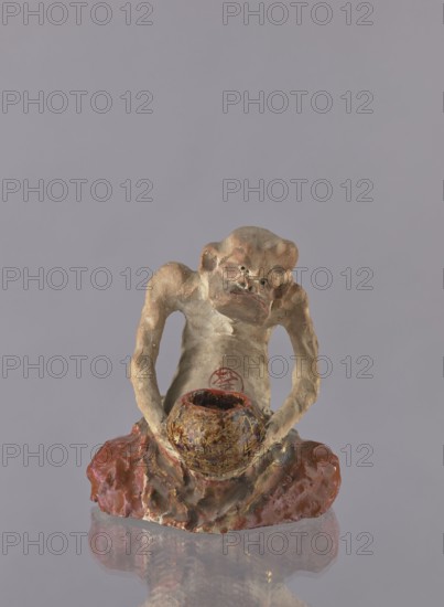 Figurine of a rakan Buddhist saint, ceramist, maker name: Choynowski, Józef 1833–ca.1915 author, Dawidow, Aleksander Pietrowicz 1838–1885 revindication, Japanese art; Japanese culture; Meiji 1868–1912; ceramics; figurines; male figurines; bowls; Buddhism; arhat iconography; rakan iconography; beggars; 1882 Meiji 15 ; Japan; sculpture; height 21.5 cm; width 19.0 cm, A small sculpted figure with a rough, textured surface in shades of beige, brown, and tan sits on a glossy reddish-brown base; the figure has an exaggerated facial expression with deep-set features and is holding a rounded, mottled bowl that shows dark brown and gold tones, while the background is a uniform light grayish-lavender color.