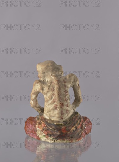 Figurine of a rakan Buddhist saint, unknown ceramist; Japanese; Meiji period 1868–1912; ceramics; figurine of a male arhat/rakan, depicted as a beggar; dated 1882 ? Meiji 15; sculpture; height 21.5 cm, width 19.0 cm., The image shows the back view of a small sculpture of a seated figure with a textured, weathered surface in shades of beige and tan, with a few areas of darker brown patina. The figure's lower garment and base display swirls of red, pink, green, and muted yellow, and there is a glossy reflection of these colors on the surface beneath it. The background is a flat, pale greyish-lavender color.